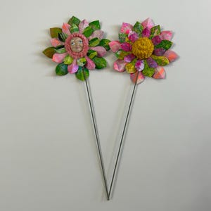 May include: Two paper flowers on wire stems. One flower has a pink and green paper design with a face in the center. The other flower has a yellow center with pink and green paper petals.