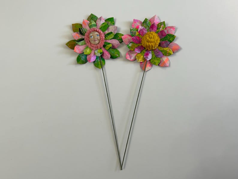 May include: Two paper flowers on wire stems. One flower has a pink and green paper design with a face in the center. The other flower has a yellow center with pink and green paper petals.