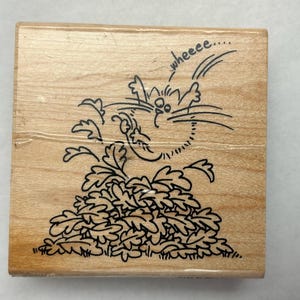 May include: Wooden stamp featuring a cartoon cat with the word "wheeeeee" above its head. The cat is drawn in black ink, with a surprised expression, and is positioned above a pile of leaves.