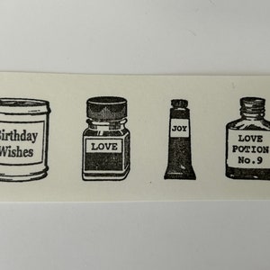 May include: Four black and white rubber stamps with different designs. The first stamp is a can with the text "Birthday Wishes". The second stamp is a bottle with the text "LOVE". The third stamp is a tube with the text "JOY". The fourth stamp is a bottle with the text "LOVE POTION No. 9".