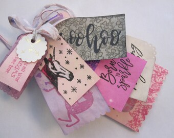 May include: A stack of pink and white gift tags with various designs, including a unicorn, a floral pattern, and the words "Born to Sparkle". The tags are tied together with a white ribbon and a small gold tag with arrows.