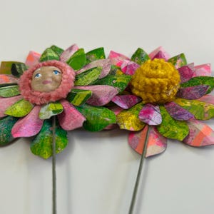May include: Two paper flowers with pink, green, and purple petals. One flower has a pink and white face in the center, and the other has a yellow pom-pom in the center.