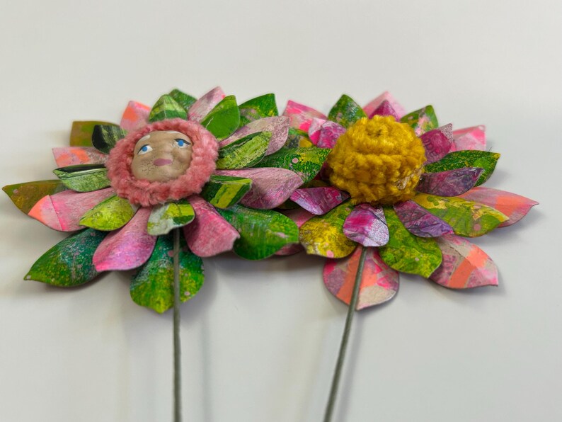 May include: Two paper flowers with pink, green, and purple petals. One flower has a pink and white face in the center, and the other has a yellow pom-pom in the center.