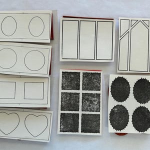 May include: A collection of rubber stamps with various designs, including circles, squares, hearts, and textured patterns. The stamps are mounted on red blocks and arranged on a white surface.