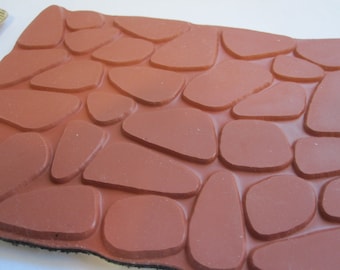 May include: A close-up of a red rubber stamp with a raised design of a cobblestone path. The stamp is designed for creating a realistic cobblestone effect on crafts and projects.