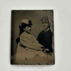May include: A vintage sepia-toned photograph featuring two people. One person is seated, wearing a light-colored coat and a hat with a decorative trim. The other person, standing behind, is wearing a dark uniform with a hat. The image has a slightly distressed appearance.