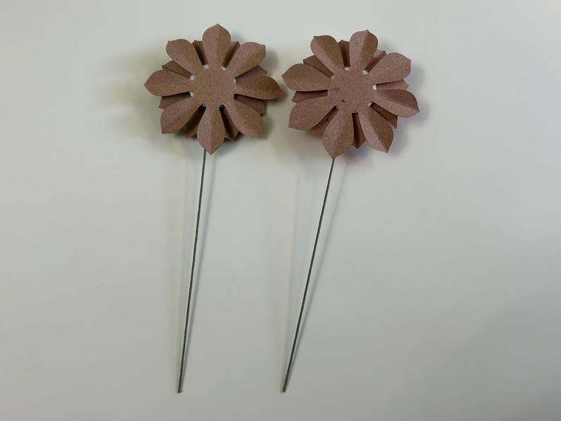 May include: Two brown paper flowers on wire stems. The flowers have a circular center and eight petals.