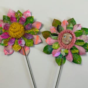 May include: Two paper flowers with green and pink petals and a yellow and pink center. The center of one flower has a painted face.