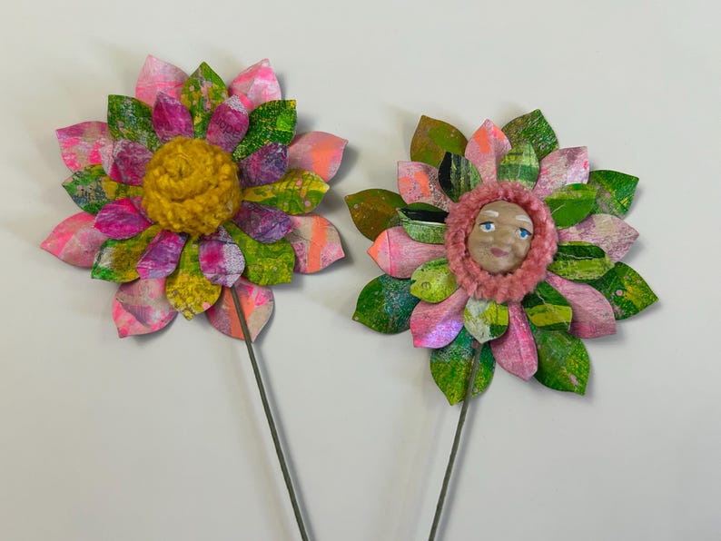 May include: Two paper flowers with green and pink petals and a yellow and pink center. The center of one flower has a painted face.