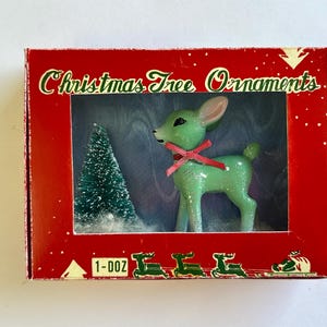 May include: A vintage Christmas tree ornament set in a red box. The box features a green deer with a pink bow and a small green Christmas tree. The box reads "Christmas Tree Ornaments".
