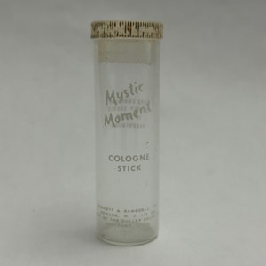 May include: A clear glass bottle with a white plastic lid. The bottle is labeled "Mystic Moment Cologne Stick" and has the text "Made in the U.S.A. by The Fuller Brush Co. Hartford, Conn." on the bottom.