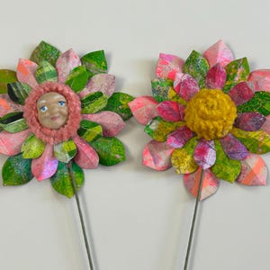 May include: Two paper flowers with pink and green petals. One flower has a face in the center and the other has a yellow pom-pom.