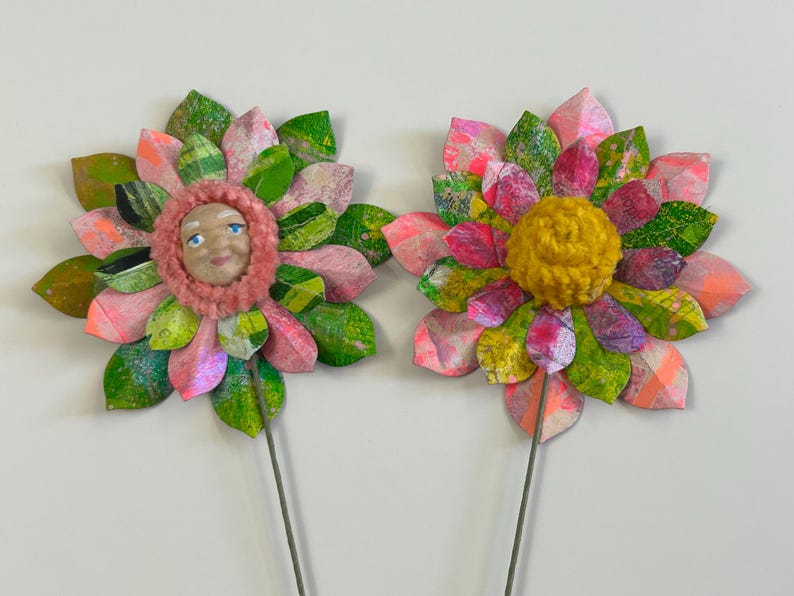 May include: Two paper flowers with pink and green petals. One flower has a face in the center and the other has a yellow pom-pom.