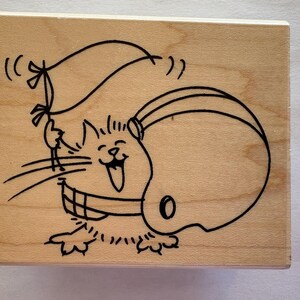 May include: A wooden stamp featuring a cartoon cat wearing a football helmet and holding a flag. The cat has a wide, happy grin and is drawn in black ink. The stamp is rectangular.