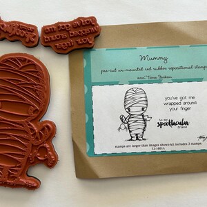 May include: A set of red rubber stamps featuring a mummy design, with additional stamps bearing text. The packaging includes a mummy illustration and the phrase "you've got me wrapped around your finger."