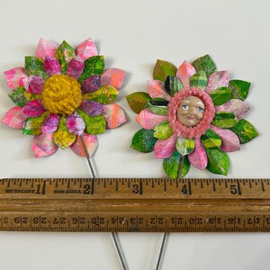 May include: Two handmade paper flowers with pink and green petals. One flower has a yellow center and the other has a pink center with a painted face.