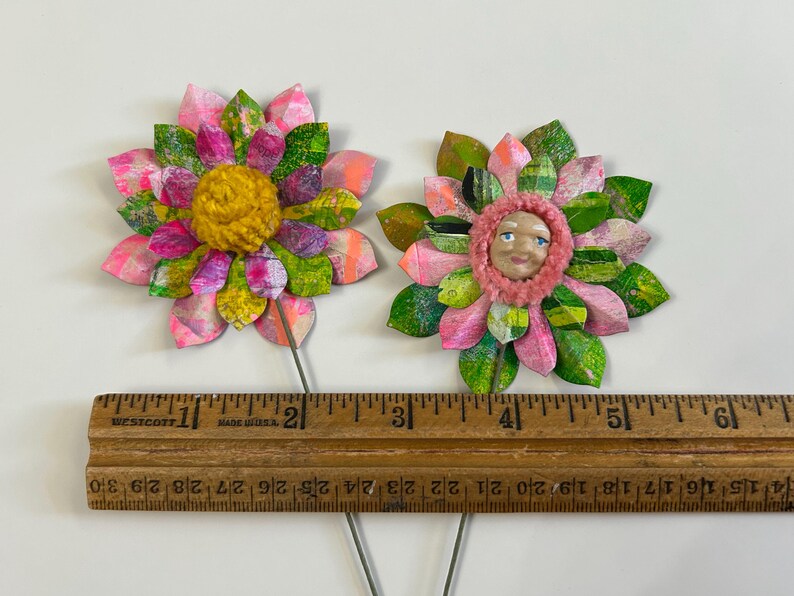 May include: Two handmade paper flowers with pink and green petals. One flower has a yellow center and the other has a pink center with a painted face.