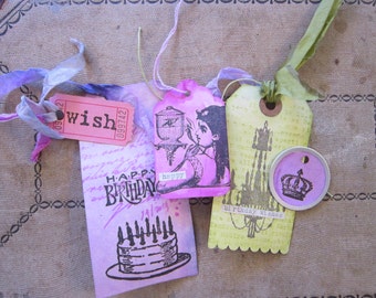 May include: A collection of decorative paper tags and a circular metal charm. The items feature printed graphics including a birthday cake, a person, a chandelier, and a crown. Text includes "wish", "HAPPY BIRTHDAY", "happy", and "birthday wishes".