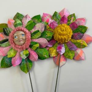 May include: Two paper flowers with pink and green petals. One flower has a yellow yarn center and the other has a pink yarn center with a painted face.