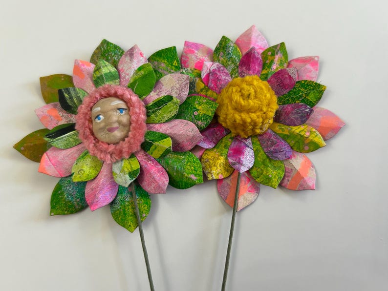 May include: Two paper flowers with pink and green petals. One flower has a yellow yarn center and the other has a pink yarn center with a painted face.