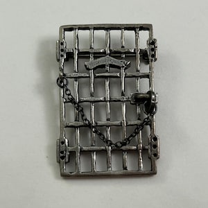 May include: A rectangular silver-toned metal brooch shaped like a prison cell door with vertical and horizontal bars. The design includes a small attached metal chain draped across the front and a central horizontal bar element with a latch mechanism.