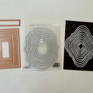 May include: A set of copper-colored rectangular metal cutting dies, a translucent plastic sleeve labeled FRAMELITS - LABELS COLLECTION (125598) containing silver-colored ornate label dies, and a black sheet with silver-colored ornate label dies.