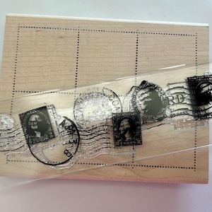 May include: A clear acrylic stamp with various vintage postage stamp designs, including portraits and text, is placed over a wooden block with dotted grid lines. The stamps feature black ink details and are arranged in a row.