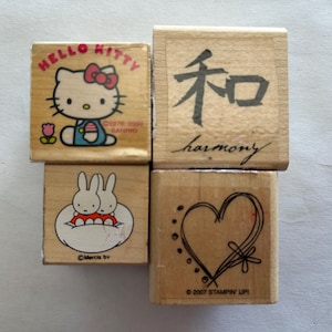 May include: Four wooden stamps with various designs. One features Hello Kitty, another has a Japanese character with the word "harmony" below it, a third shows two bunnies, and the last has a heart design.
