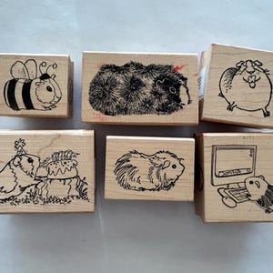 May include: Six wooden stamps featuring illustrations of guinea pigs. The stamps depict a guinea pig with a bee, a guinea pig with a birthday cake, a guinea pig with a computer, and three different guinea pigs.