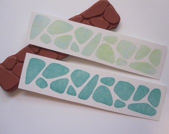 May include: Two white cards with a pattern of green and blue stones printed on them. The stones are different sizes and shapes. The cards are next to a brown rubber stamp with a similar stone pattern.