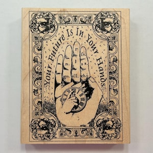 May include: Wooden stamp with a hand illustration and the text "Your Future Is In Your Hands." The design includes a clock, eyes, and decorative borders. The stamp is light brown with black ink.