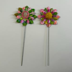 May include: Two colorful paper flowers on wire stems. The flower on the left has a pink and green color scheme with a face in the center. The flower on the right has a pink, green, and yellow color scheme with a yellow center.