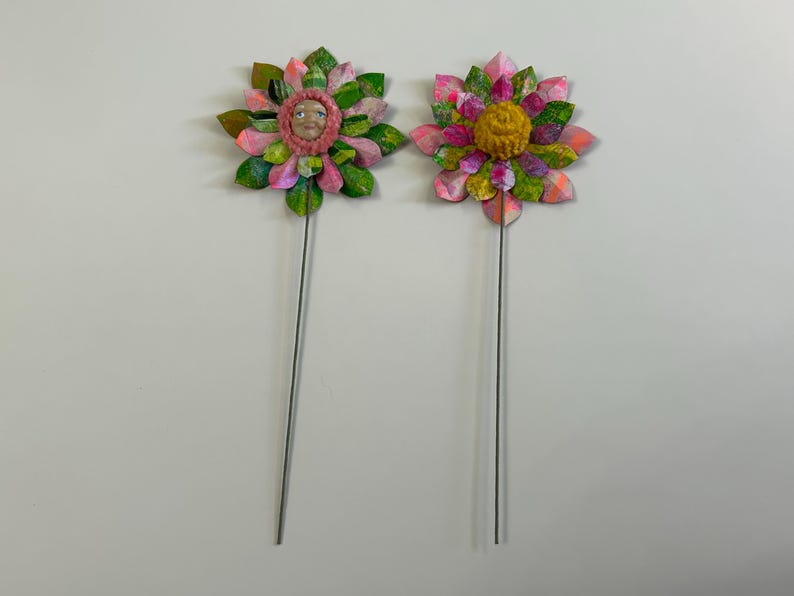 May include: Two colorful paper flowers on wire stems. The flower on the left has a pink and green color scheme with a face in the center. The flower on the right has a pink, green, and yellow color scheme with a yellow center.