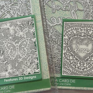 May include: Two card dies with intricate designs. One features butterflies, the other a heart with the words "lot of love." Both are white cut-outs on a green background. The packaging reads "Features 3D Designs" and "Craft a Card Die."