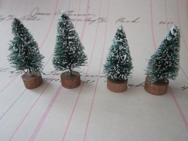 Peut inclure: Quatre mini sapins de No&euml;l artificiels avec des aiguilles vertes et un flocage blanc, chacun sur un socle en bois brun.