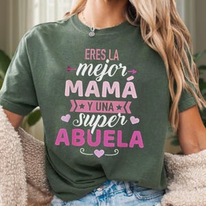 May include: A dark green t-shirt with the Spanish text "Eres la mejor mamá y una super abuela" in pink. The text is arranged in a layered design with decorative arrows, stars, and hearts. The shirt is a crew neck style.