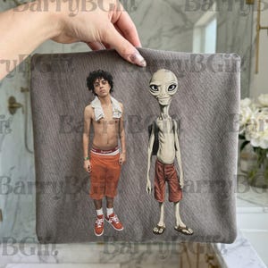 May include: Gray rectangular pillow featuring a graphic of a person and an alien. The person is wearing orange shorts, red sneakers, and a towel. The alien is wearing red shorts and sandals.