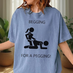 May include: Light blue t-shirt with the words "BEGGING" above a black graphic of two stick figures in a sexual position, and the words "FOR A PEGGING!" below the graphic. The shirt is made of a soft material.