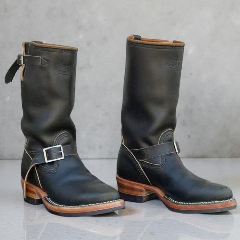 May include: A pair of black leather boots with a mid-calf height. The boots feature a buckle strap, a wooden heel, and a light-colored sole. The upper part of the boot is smooth, and the heel is approximately 2 inches high.