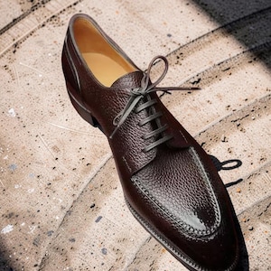 Peut inclure: Une chaussure de ville en cuir marron avec des lacets gris. La chaussure a une surface texturée et un design cousu autour de l'orteil. La chaussure est de taille 43.