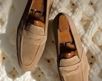 May include: A pair of tan suede penny loafers with wooden shoe trees inside. The shoes have a classic design with a rounded toe and a strap across the vamp. The stitching is visible, and the shoes are set on a white, textured surface.