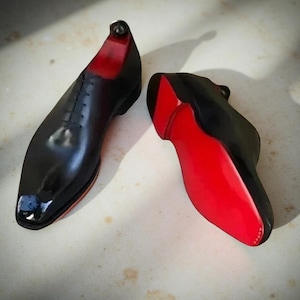 Op de afbeelding: Paar zwarte leren herenschoenen met rode zolen. De schoenen hebben een klassiek vetersluiting ontwerp en een glanzende afwerking. De rode zolen contrasteren met het zwarte bovenwerk.