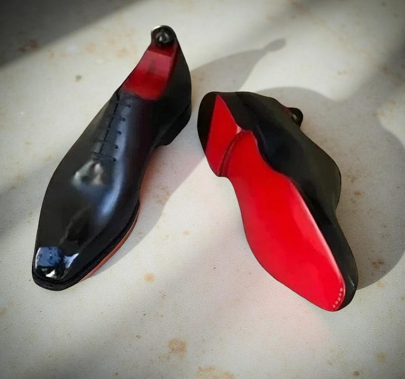 Op de afbeelding: Paar zwarte leren herenschoenen met rode zolen. De schoenen hebben een klassiek vetersluiting ontwerp en een glanzende afwerking. De rode zolen contrasteren met het zwarte bovenwerk.