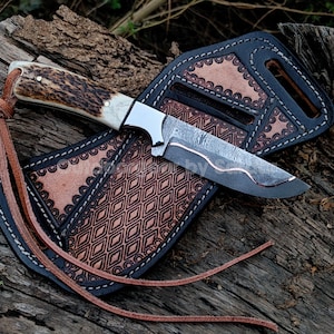 Könnte beinhalten: Ein feststehendes Messer mit einem strukturierten Hirschhorngriff und einer Damaststahlklinge mit wellenförmiger Kupfereinlage. Dazu gehört eine braune Lederscheide mit schwarzem Rand, aufwendiger geometrischer Prägung und einem Lederband.