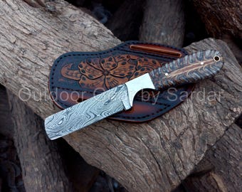May include: A handcrafted knife with a patterned blade and a wooden handle rests on a brown leather sheath with a floral design. The knife is set against a textured wooden background. The text "Outdoor Gear by Sardar" is visible.