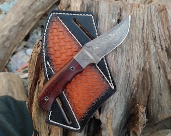 May include: A small, brown-handled knife with a patterned blade, in a black leather sheath with an orange stitched design.