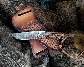 Peut inclure: Un couteau de chasse artisanal avec une lame à motifs et un manche décoratif, reposant dans un étui en cuir marron. L'étui présente un motif tissé et une lanière en cuir. Le couteau et l'étui sont présentés sur une surface en bois.