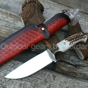 Peut inclure: Un couteau de chasse avec une lame en acier poli et un manche en os et en corne. Le couteau est accompagné d'un étui en cuir rouge avec un motif tissé et une lanière en cuir. Le texte "Outdoor gears by ordar" est visible.