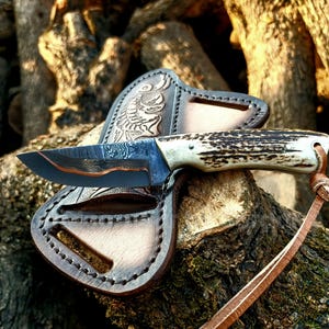 Peut inclure: Un couteau de chasse artisanal avec une lame en acier à motifs et un manche en os et en bois. Le couteau repose sur un étui en cuir marron avec des gravures décoratives. Une lanière en cuir est attachée au manche.