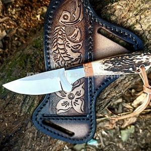 Peut inclure: Un couteau de chasse artisanal avec une lame en acier poli et un manche en corne. Il est présenté dans un étui en cuir personnalisé avec des motifs floraux et de poisson en relief. L'étui est marron foncé avec des coutures noires.